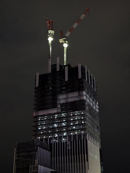 At night, two white-red cranes stand tall on a skyscraper under construction. Their supports flood lamps pointing downards, illuminating their supports. Many unfinished floors are being lit up from the inside, visible through the not yet closed off building hull.