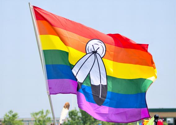A brightly colored 2-Spirit Pride flag ripples on a metal pole against a clear pale sky. Seven horizontal rainbow stripes (red, orange, yellow, green, blue, indigo, violet) bear a central white circle enclosing a stylized feather emblem. A single real feather is tucked into the pole near its base.