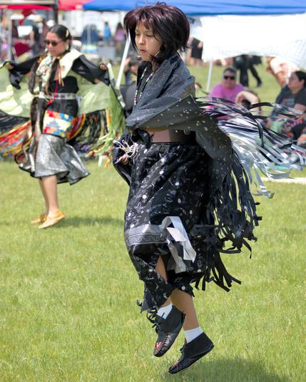 A dancer leaps in mid-step on green grass, wearing a black shawl patterned with tiny silver stars and long black-and-silver ribbon fringe that streams outward. The matching black skirt also features a star and planet print, and the dancer wears white ankle socks and black leather moccasins. 