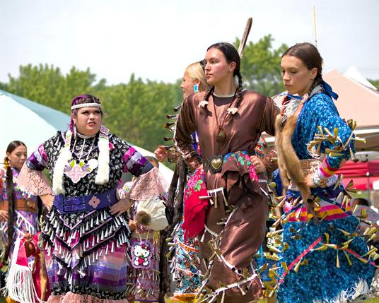  Three jingle dancers stand in a row before brightly colored canopies. From left: one wears a black, purple and pink jingle dress with layered bell fringes and white fur accents; the center dancer wears a brown dress with a heart-shaped belt buckle, jingles at wrist and waist level, and carries a red handkerchief; the right dancer wears a blue floral dress with yellow cone bells and fur trim. All have feathers in their hair and hands on hips, poised to dance.
