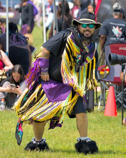 A smiling dancer leans forward in dance on a grassy field, wearing a black wide-brimmed hat trimmed with beads, mirrored green sunglasses, a purple-and-yellow fringed shawl over a black shirt, a purple sash at waist level, and jingle-bell ankle cuffs over black fur mukluks. 