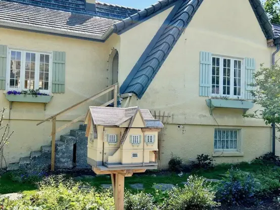 The image features a charming yellow house with a blue-tiled roof and green shutters on the windows. The house has a small staircase leading up to the entrance, with a wooden handrail. In the foreground, there is a large, detailed wooden birdhouse designed to resemble a miniature house, complete with a gray roof and white trim, mounted on a wooden post. The garden in front of the house is well-maintained, with various plants and flowers, including purple flowers in the window boxes. The scene i…