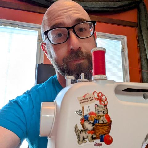 A photo of me, a 47 year old white man with a shaved head, a short cropped brown & grey beard, 

photographed from the chest up,

wearing a bright blue T-shirt and black acetate glasses

behind a sewing machine with red thread on the spool and a sticker of cute kittens in a basket with yarn and a slogan reading "Sewing is a radical act"