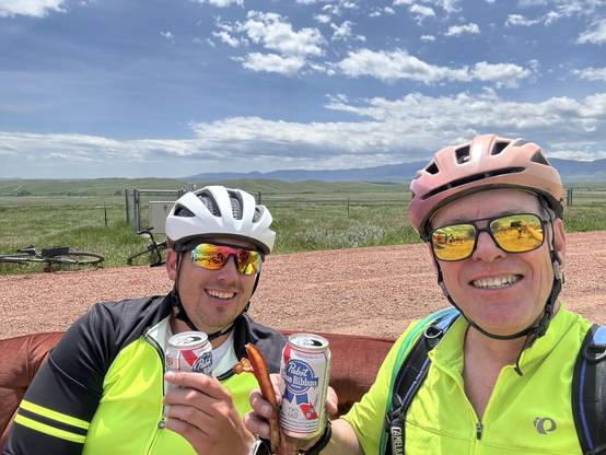 Two happy gravel racers holding cans of Pabst beer and strips of bacon under sunny Wyoming skies