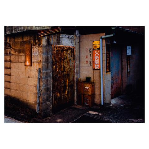 A narrow alley corner shows the side of a small, weather-worn industrial building. The cinder block walls are stained and spotted with rust. Several metal signs warn against fire (火気厳禁), though the corroded state of the doors suggests that enforcement is optional. Two metal cabinets, each battered and tinged with rust, sit outside near a tiny potted plant—nature’s lone attempt at decor.