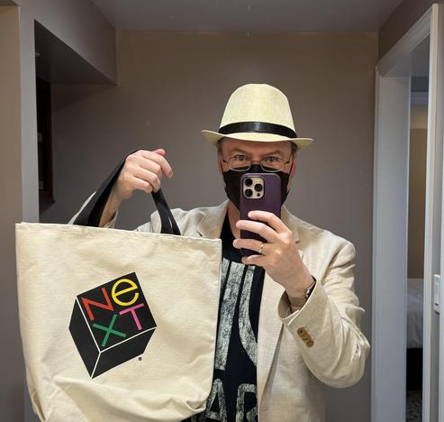Me, wearing a black mask, a light fedora, and a light linen jacket over a black Atari t-shirt, holding up a NeXT tote bag.
