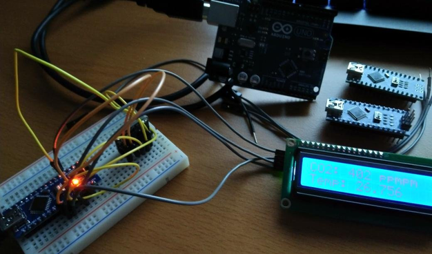 **TL;DR:** Image shows a CO₂ monitoring setup in progress (AirMaster3000), using Arduino components, a breadboard, sensors, and an LCD screen displaying data.

**Extracted Text:**
CO2: 402 PPM
Temp: 26.756

**Description:**
On a wooden desk is a DIY electronics project labeled "AirMaster3000", a CO₂ measuring device. A small breadboard has an Arduino Nano connected with several colored jumper wires, and an orange LED is lit on the board. The setup includes a larger Arduino Uno connected via USB, and two unused Arduino Nano boards placed nearby. A green-framed LCD screen shows live data: "CO2: 402 PPM" and "Temp: 26.756". The device is mid-development and displays real-time air quality and temperature readings.
