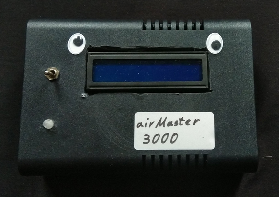 **TL;DR:** Final build of the AirMaster3000 CO₂ sensor in a black case, labeled and humorously decorated with googly eyes.

**Extracted Text:**
**airMaster 3000**

**Description:**
This image shows the fully enclosed AirMaster3000 CO₂ measuring device in a matte black plastic case. It features a central blue LCD screen, a metal toggle switch, a small white indicator light or sensor, and a handwritten label that reads “airMaster 3000.” Two googly eyes are attached above the screen, giving the device a fun, anthropomorphic look—resembling a little robot face. The case has air vents, likely for sensor airflow.
