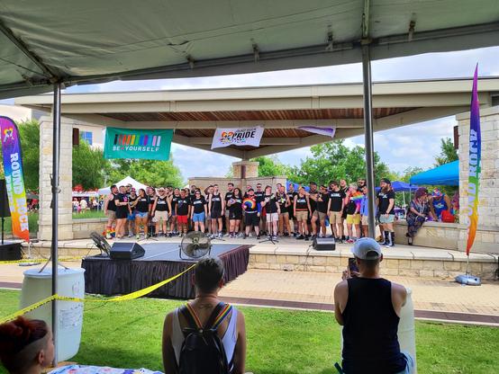 Photo of Austin Gay Men's Chorus, around 30 people in black AGMC t-shirts and colorful shorts, singing on stage in an outdoor ampitheater, at the Round Rock Pride Fest, Texas 6/7/2025