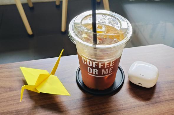This photo shows a wooden table with three items on it. In the center is a clear plastic cup with a dome lid and black straw, containing what looks like iced coffee - it has layers of brown coffee and lighter cream or milk. The cup has text that reads "COFFEE OR ME" in white letters. To the left of the cup is a small yellow paper origami crane. To the right is a white rounded object that appears to be wireless earbuds or a small electronic device. The table appears to be in a café setting with wooden chairs visible in the blurred background. [by claude ai]