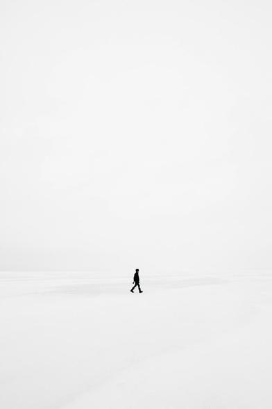 A solitary figure walks across a vast, empty landscape of white or light gray terrain under an overcast sky, creating a minimalist composition that emphasizes isolation and the scale of the surrounding space.​​​​​​​​​​​​​​​​ Image shared in 8 June 2025 Home Base with Jeff Warren Newsletter.