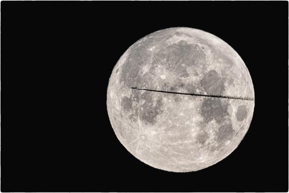 Schwarzweissfotografie des Vollmondes, vor dem ein Flugzeug mit Kondensstreifen vorbeifliegt. 