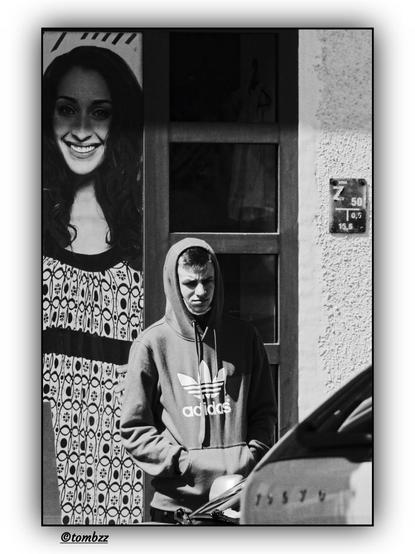 A young boy in a gray Adidas hoodie stands in front of a window, looking down. His face has a serious, focused expression. Behind him, a billboard features a young woman in a black-and-white dress—her face radiates a joyful, wide smile, creating a striking contrast with the boy's stern expression. 
The composition of the photograph highlights the emotional disparity between the boy's reality and the idealized happiness presented on the billboard, sparking reflection on the contrast between advertising imagery and real human emotions on the street.