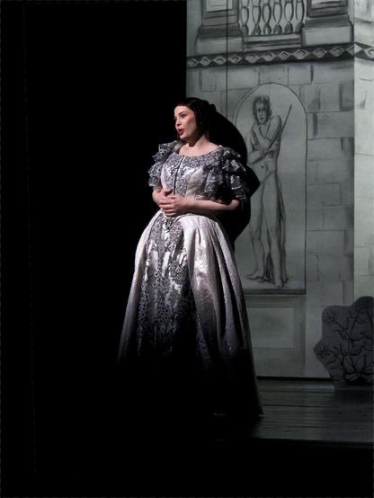 Woman with dark hair wearing a white dress decorated with black lace down the center stands poised, her hands gently clasped. In the background is a white stage set showing classical architectural elements and a male statue drawn in black. Soprano Megan Marie Hart as Elvire in Paul Georg Dittrich's staging of Daniel Auber's grand opéra « La Muette de Portici » at Staatstheater Darmstadt, 2025. photo: © Yan Melo Hoffmann-Rothe