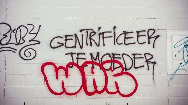 A cinder block wall painted white, words "GENTRIFICEER JE MOEDER"* spray painted on it in black. "WAR" spray painted below in red below, something that looks like artist signature is spray painted in thinner black to the left of the main message.

* from Dutch: "gentrificate your mother"