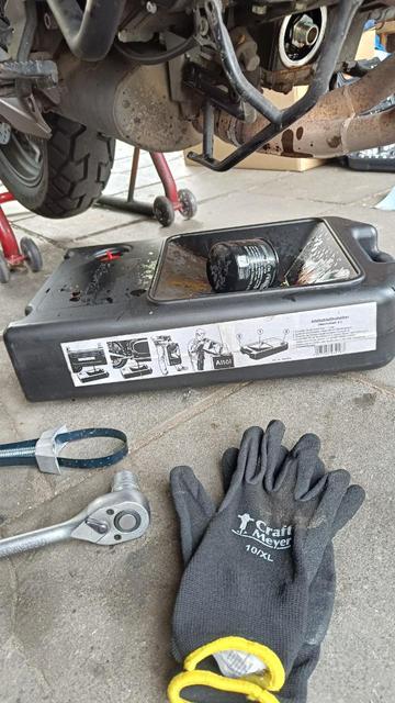 Underside of a motorcycle, with an oil drain pan under it. The spent oil filter lies in the drain pan, some tools and gloves in the foreground.