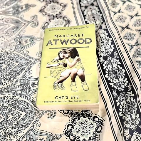 Alt-text:
Paperback copy of Margaret Atwood's "Cat's Eye" resting on layered vintage lace and patterned fabrics. The yellow book cover features two girls in summer dresses sitting close together, with "Shortlisted for the Man Booker Prize" noted below the title.


Secondary Description:
Like an archaeological discovery, Atwood's Cat's Eye emerges from layers of intricate lace—delicate paisley spirals and floral motifs creating a textile palimpsest beneath the book's weathered spine. The cover's two girls, frozen in their childhood intimacy, seem to whisper secrets across the ornate fabrics that cradle them. One blonde, one brunette, they sit in eternal summer yellow, their small bodies pressed together in that particular closeness that precedes betrayal or transcendence.


The decorative patterns beneath—swirling paisleys like fossilised tears, geometric flowers blooming in black and white—create a mandala of memory, each thread a story within the story. The book rests here not merely as an object but as a portal; its Man Booker Prize notation is a quiet testament to the power of bearing witness. In this tableau, domestic beauty and literary weight converge, the delicate lacework beneath echoing the novel's intricate exploration of girlhood's sharp-edged beauty—all surfaces and hidden depths, patterns that conceal and reveal the truths we carry.
