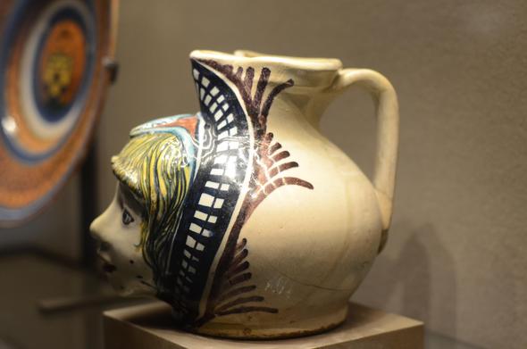 A wide pitcher glazed in many colors, with the face of a boy sculpted on the belly. He's got yellow hair and a bit of a panicky look on his face.  