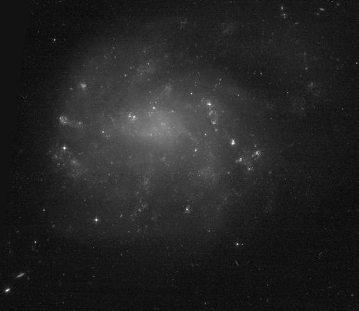 A greyscale image of a spiral galaxy. The faint galaxy has a central bar and loose spiral arms that are not very well defined, which arc clockwise around the bar. The arms are dotted with bright star clusters.