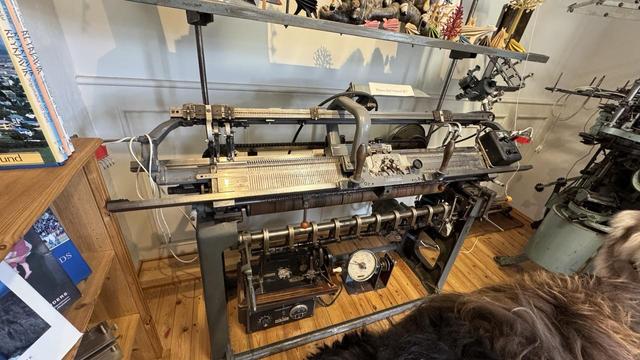 A vintage motorized v-bed knitting machine. It has a fair number of patterning and takedown widgets such that it could be used for fairly automatic production. There are craft-shop knickknacks are books around it. This was in Alafoss yarn shop, and was not currently in use — seemed to be there for decorative purposes — but it looked like it was in operating condition. 