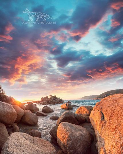 A dramatic coastal scene from Wilsons Promontory at sunset. Warm golden light from the low sun illuminates smooth, rounded granite boulders in the foreground as waves gently flow around them. In the midground, more rocks extend into the sea, crowned by a small tree. The sky above is painted with striking purples, oranges, and blues, with textured clouds catching the fading light. Distant hills on the right complete the tranquil composition.