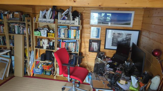 Interior of the corner of the shed where I mostly 'work'. Wood horizontal planks with log cabin butt joints. 

Pictures on the walls, a stretch of 4 bookcases along the wall my red spinny chair and the glass table obscured by stuff, the detritus and tools of a computer person. 

A new jute or sizal woven mat edged in red which replaces the previous one without the ripped edging and dingier weave.