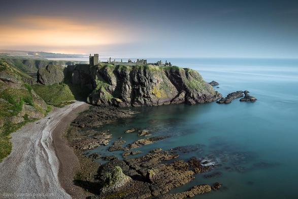 Dunnottar Castle is bathed in a soft, silvery glow under the moonlight. The ancient stone structure stands majestically on a rugged cliff, surrounded by a tranquil sea. In the distance, the lights of Aberdeen are visible on the horizon. The landscape is serene, with the moon casting gentle shadows and highlighting the castle's historic features against the night sky.