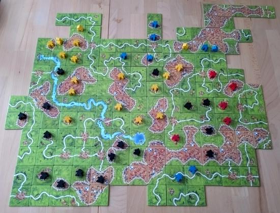 Holztisch auf dem das fertig gespielte Spiel Carcassonne liegt. Zu sehen sind Plättchen auf denen ein Fluss zusammengelegt würde, Burgen, Straßen mit unterschiedlich farbigen Spielfiguren darauf. So ist eine individuelle Landschaft entstanden.