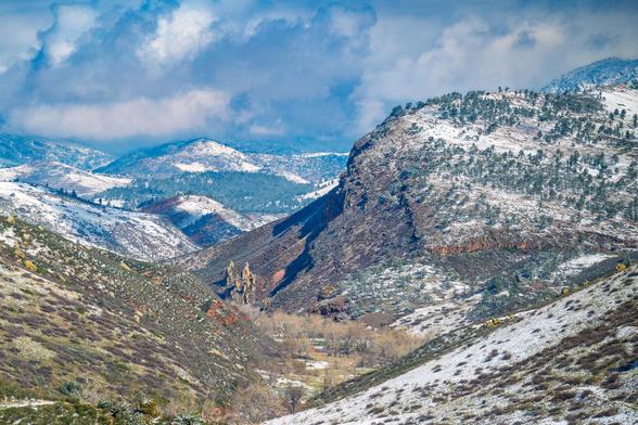 Landscape photo of Rabbit Mountain Open Space in Colorado after fresh snowfall. The canyon and surrounding ridgelines are partially dusted with snow, with patches of green trees and red rock formations visible beneath a dramatic sky.