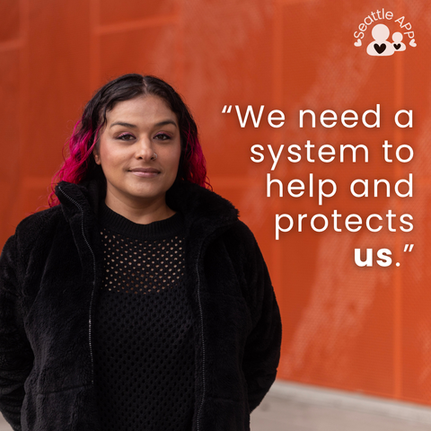 Image description: Image is of a woman looking into the camera and smiling with her hands behind her back. In the corner of the image is the Seattle APP logo.

On top of the photo is a transparent black box with white lettering. The text in the middle of the box is the following “We need a system to help and protects us.”

