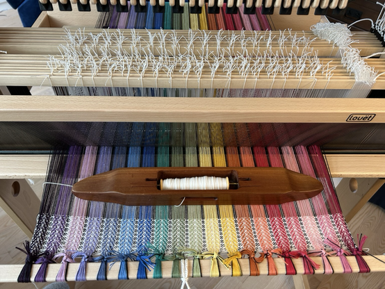 A view of a handloom with a hand carved wooden boat shuttle sitting on top of a warp in rainbow coloured stripes with black stripes between each colour band 