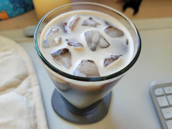 Clear pint glass filled to the top with iced Kirkland Signature Colombian Cold Brew Coffee, on a gray diatomaceous-earth coaster on an off-white desk mat on a maple desk. The top has foamed coffee and it looks delicious.
