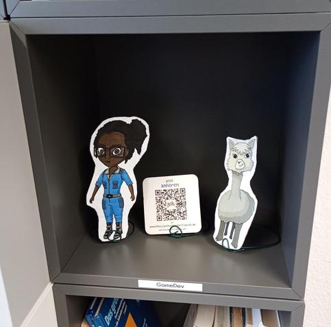 Bild eines grauen IKEA Eket-Regalwürfels im Hackerspace Bremen e.V. mit der Beschriftung "GameDev", in dem sich drei Pappaufsteller befinden. Zwei davon sind Spielcharaktere aus einem Raumschiff-Spiel, der dritte macht Werbung für HobbySpieleentwicklerPodcast.de.