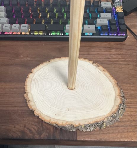 On a desk sits a DIY DVD holder made of a wooden dowel rod inset into a slice of tree trunk. The materials are made in USA and purchased from Michaels craft store. The cost is cheaper than a plastic equivalent if you already own a drill.