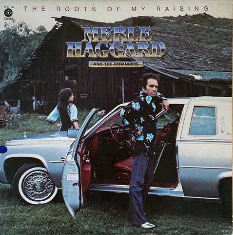 album cover for Merle Haggard's 1976 "the Roots of my Raising", with a photo of him standing in the open door of a Cadillac(?). a woman with long dark hair faces away. the surrounding scenery is a rural setting with an old farmhouse in the background.
the album's title is written in a simple sans serif along the top while the artist's name is, as the post says, capitalized serif desert chrome.