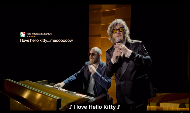 Two men perform on stage, one playing a keyboard while the other sings. They wear sunglasses and appear to be having fun. Above them is a app review referencing "Hello Kitty." The setting features warm lighting and a backdrop of wooden panels.
