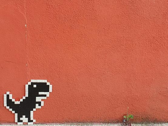Picture of a plastic mosaic of a pixelated T-Rex in black with a white contour, stuck on a red wall at ground level. There is a small green plant growing further away at the bottom of the wall. The T-Rex picture is the one from the Chrome browser mini-game.