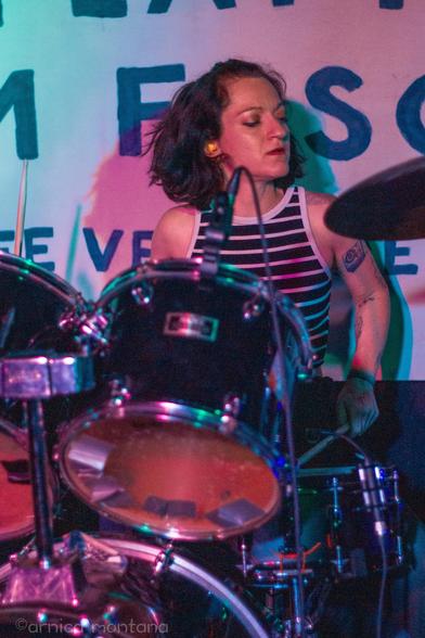 A woman with short, dark hair plays the drums on stage, wearing a striped tank top. The setting is vibrant, illuminated with colorful lights, and a partially visible backdrop displays text.