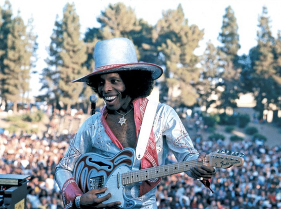 Sly on an outside stage, wearing a silver outfit and matching hat, with the crowd behind him.