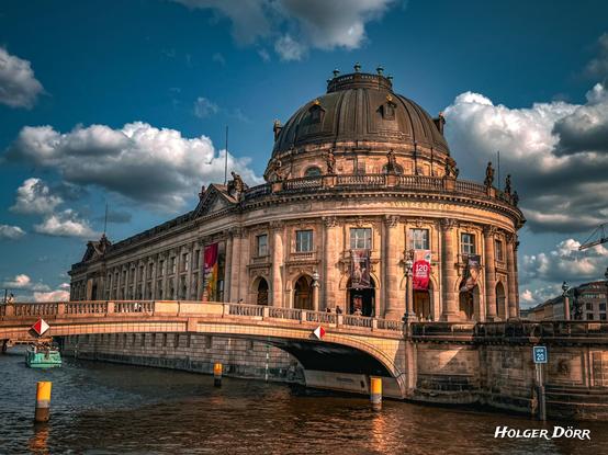 Das Bode-Museum in Berlin im goldenen Abendlicht, aufgenommen von der Brücke an der Spree. Die markante Kuppel leuchtet warm vor einem teils wolkigen Himmel.