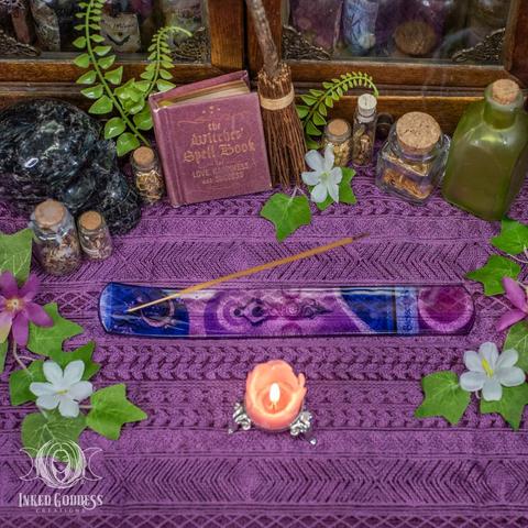 An example of the Spiral Goddess Glass Incense Holder sits on a purple lace-covered surface. It's a long flat rectangle with rounded corners, and one end is slightly bent up to hold the incense stick above the rest of the plate. It's printed with crescent moons, triple moons, and a spiral goddess design. Presented by Inked Goddess Creations.