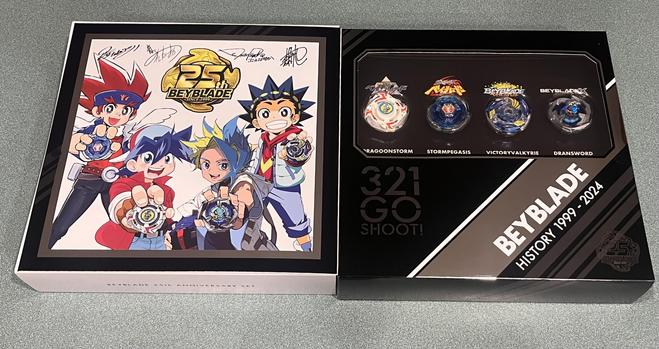The Beyblade 25th Anniversary box opened showing the series’s four protagonists holding their Beys with signatures above them on the left, and on the right it says “3 2 1 GO SHOOT!” and “BEYBLADE HISTORY 1999 - 2024” below a window showing BEYBLADE X versions of Dragoon Storm, Storm Pegasus, and Victory Valkyrie, as well as a version of DranSword with holographic sticker.
