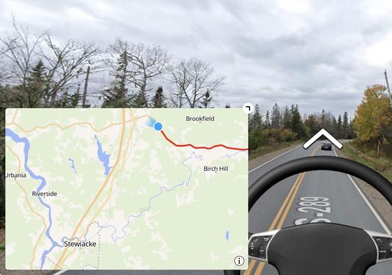 Alt text: A simulated driving view on Highway 289 in Nova Scotia, with a steering wheel in the foreground and a map overlay on the left showing a route from Brookfield toward Stewiacke. The GPS marker indicates the current location near Brookfield. The surrounding area features a tree-lined rural road under a cloudy sky. Destination: Stewiacke, to see a mastodon exhibit.