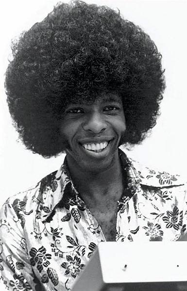 A black and white photograph of a youngish Sky Stone smiling beautifully with giant lamb chops and an incredible afro, wearing an embroidered floral shirt.