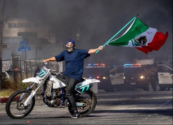 Motorcyclist holding the mexican flag blowing in the wind while police are in the background surrounded by black smoke  