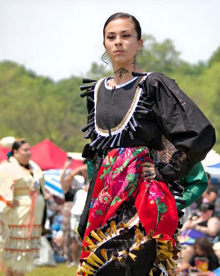 A three-quarter portrait of a jingle-dress dancer poised on a grassy powwow arena. The dancer has sleek hair pulled back, small tattoos of dots above each eyebrow, and a stylized bird motif tattooed on the throat. They wear a black satin dress trimmed with lace and rows of black metal jingles around the yoke and skirt. A red floral shawl is knotted at the waist, and their right hand rests on the hip. In soft focus behind are other dancers and festival spectators under colorful canopies.