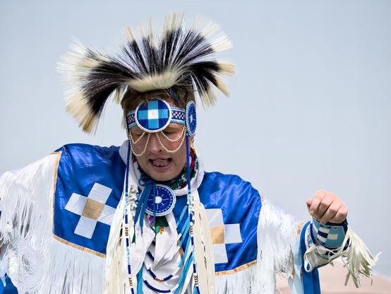 A mid-shot of a powwow dancer captured outdoors against a pale blue sky.  A beaded headband in a blue-and-white checkered pattern crosses the forehead, with two round medallions over each temple linked by gold-tone bead chains that drape across the face. Atop the head sits a tall black-and-white porcupine-hair roach. The dancer’s eyes are closed, mouth slightly open with tongue visible, and arms are lifted in mid-motion.