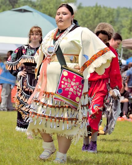 A full-body view of a jingle-dress dancer walking on a grassy field. The dancer wears a cream-colored dress covered in rows of silver cone-shaped jingles suspended from orange-and-black patterned ribbon. Scalloped cuffs on the sleeves and hem feature matching floral trim. A square red cloth bag with gold-leaf appliqués and colorful flowers hangs diagonally from the shoulder. White leggings and beaded moccasin boots complete the outfit. In soft focus behind, other dancers in dark and red regalia and spectators under canopy tents can be seen.