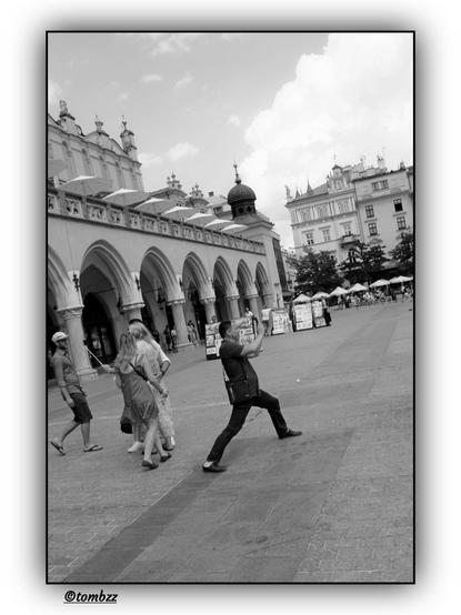 A black-and-white photograph captures a dynamic scene in the main square of Kraków. In the foreground, a man stands with a wide stance, leaning forward intensely to take the perfect shot of something outside the frame. His posture and focus convey full determination and commitment to achieving an ideal photograph.
Behind him, four people are standing. Three of them are absorbed in taking a selfie—one person is wearing a hat and shorts, another is dressed in a dress, and the third is wearing a long gown. Their relaxed poses contrast with the main subject’s professional approach to photography, creating an interesting juxtaposition between spontaneous fun and serious dedication.
In the background, a picturesque view of historic buildings unfolds, including the characteristic Cloth Hall with elegant arches and other architectural landmarks.