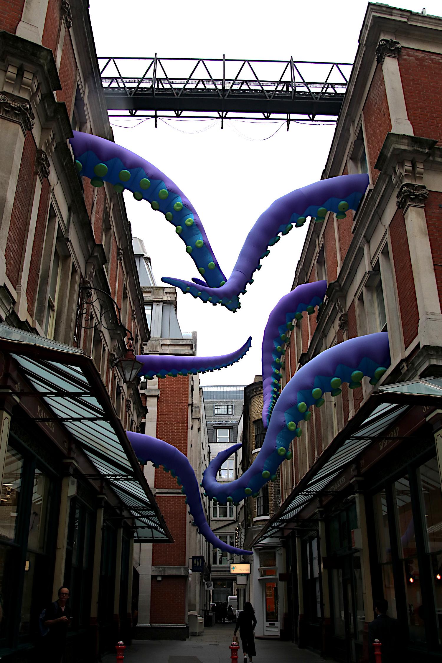 Blick in eine Passage in London. Aus den Gebäuden links und rechts ragen lila Tentakel mit Saugnäpfen.